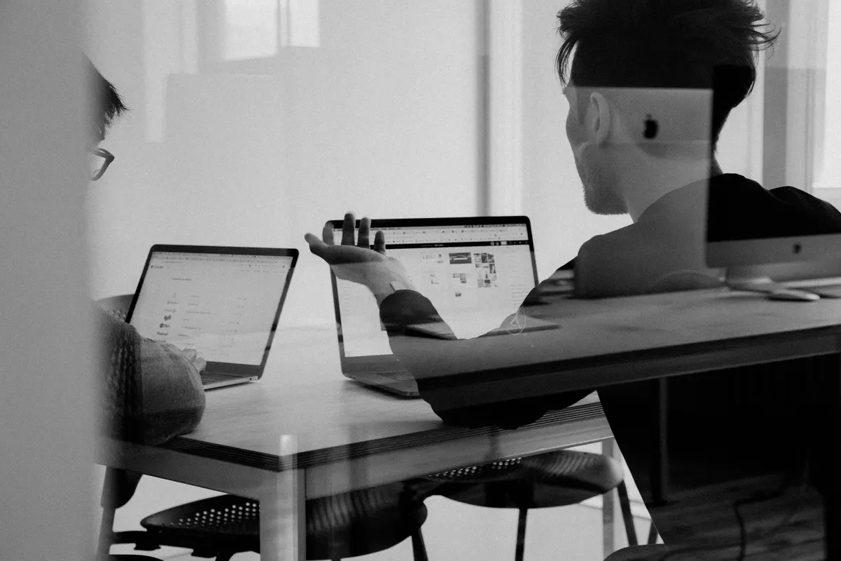 Two collaborators reviewing work on laptops through a glass meeting-room wall.