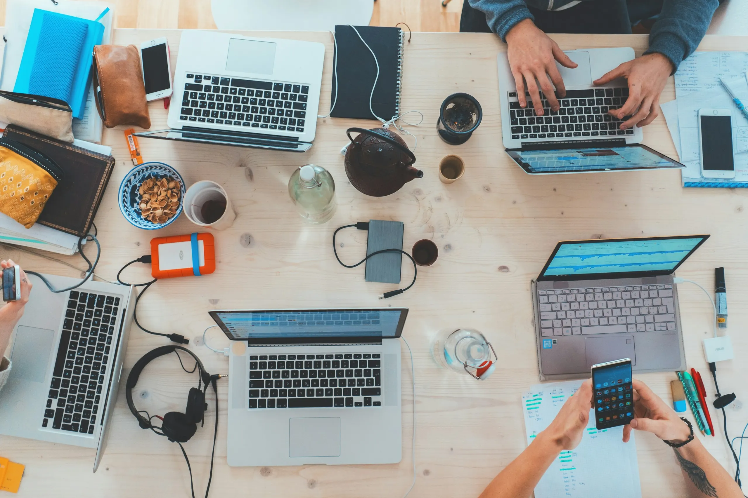 An overhead view of people using several laptops and mobile devices at a shared table.
